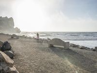 a couple are walking down the beach towards the ocean with rocks, boulders and benches