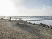 a couple are walking down the beach towards the ocean with rocks, boulders and benches