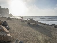 a couple are walking down the beach towards the ocean with rocks, boulders and benches