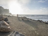 a couple are walking down the beach towards the ocean with rocks, boulders and benches