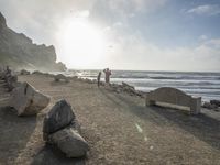 a couple are walking down the beach towards the ocean with rocks, boulders and benches