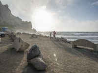 a couple are walking down the beach towards the ocean with rocks, boulders and benches