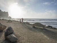 a couple are walking down the beach towards the ocean with rocks, boulders and benches