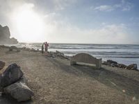 a couple are walking down the beach towards the ocean with rocks, boulders and benches