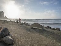 a couple are walking down the beach towards the ocean with rocks, boulders and benches