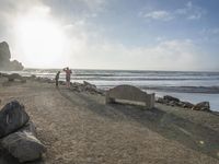 a couple are walking down the beach towards the ocean with rocks, boulders and benches