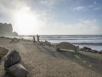 a couple are walking down the beach towards the ocean with rocks, boulders and benches