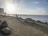 a couple are walking down the beach towards the ocean with rocks, boulders and benches