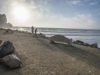 a couple are walking down the beach towards the ocean with rocks, boulders and benches