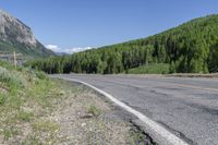 the paved road passes a wide valley with lots of green trees along it and tall mountains in the distance