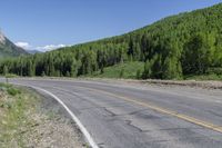 the paved road passes a wide valley with lots of green trees along it and tall mountains in the distance