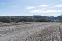 an empty gravel road on a mountain side with rolling hills in the background, and mountains in the distance