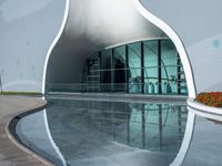 a curved building with a reflection of the water and plants around it outside of the building