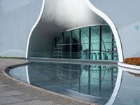 a curved building with a reflection of the water and plants around it outside of the building