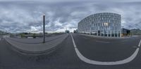 a fisheye photograph of a curved city street in the middle of cloudy day with a building and street light nearby