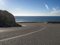 a curve of paved road with a rocky cliff above it with sea and clouds in background