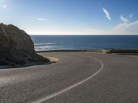 a curve of paved road with a rocky cliff above it with sea and clouds in background