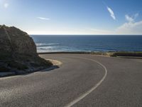 a curve of paved road with a rocky cliff above it with sea and clouds in background