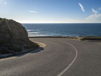 a curve of paved road with a rocky cliff above it with sea and clouds in background