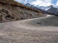 a dirt road winding in the mountains towards some snow - covered mountains and some brown rocks