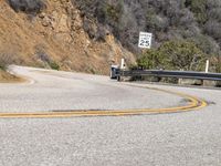 there is a sign posted to the side of a curved road near some hills or bushes