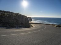 a curved curve of road next to a hill near the ocean with bright sun in the distance