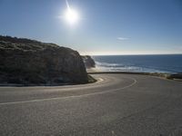 a curved curve of road next to a hill near the ocean with bright sun in the distance