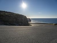 a curved curve of road next to a hill near the ocean with bright sun in the distance