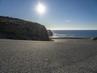 a curved curve of road next to a hill near the ocean with bright sun in the distance