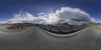 a fish eye view of a curved road near a mountain range in the desert with a cloudy sky above