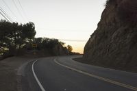 a curved roadway going next to the mountain side with power lines above it and telephone poles on the other side