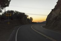 a curved roadway going next to the mountain side with power lines above it and telephone poles on the other side