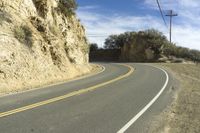 a curved roadway going next to the mountain side with power lines above it and telephone poles on the other side