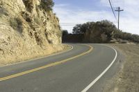 a curved roadway going next to the mountain side with power lines above it and telephone poles on the other side