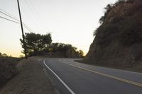 a curved roadway going next to the mountain side with power lines above it and telephone poles on the other side