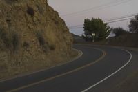a curved roadway going next to the mountain side with power lines above it and telephone poles on the other side