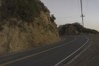 a curved roadway going next to the mountain side with power lines above it and telephone poles on the other side