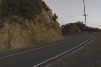 a curved roadway going next to the mountain side with power lines above it and telephone poles on the other side