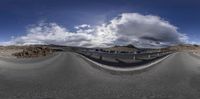 a fish eye view of a curved road near a mountain range in the desert with a cloudy sky above