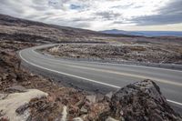 an empty curved road going between rocky and desert hills with mountain backdrop, with some rocks and a single motorcycle in the foreground