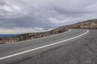 a curved road is next to a rocky hillside and ocean shore at the foot of a steep, rocky hill