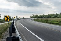 a curved road with signs on the roadsides in front of the sky with clouds and green grass