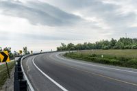 a curved road with signs on the roadsides in front of the sky with clouds and green grass