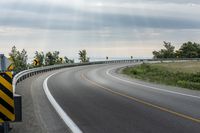 a curved road with signs on the roadsides in front of the sky with clouds and green grass