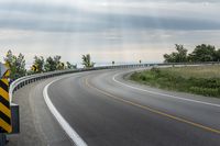 a curved road with signs on the roadsides in front of the sky with clouds and green grass