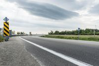 a curved road with signs on the roadsides in front of the sky with clouds and green grass