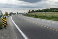 a curved road with signs on the roadsides in front of the sky with clouds and green grass