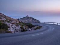 a curved roadway windscreen near the ocean and a hill top lighthouse at dusk - photo of twilight