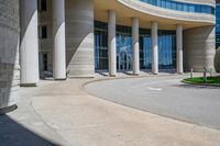 a curved sidewalk leading to an entrance of a building, with columns and bushes on each side of it