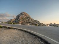 a motorcycle parked by the side of a mountain road at sunset, with a road and rocks in the background